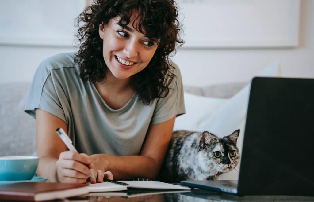 Mulher de cabelo cacheado e franja, sorri para a tela do notebook, um gato está ao seu lado. Ela escreve sobre marketing de conteúdo em um caderno.