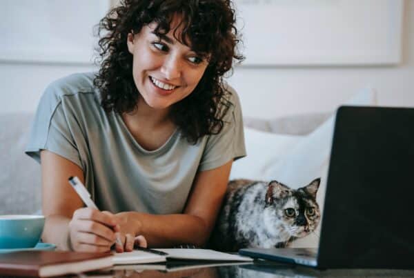 Mulher de cabelo cacheado e franja, sorri para a tela do notebook, um gato está ao seu lado. Ela escreve sobre marketing de conteúdo em um caderno.